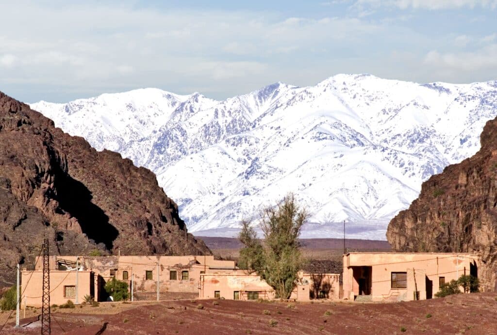 Petit village aux maisons ocre situé dans une gorge désertique, avec les sommets enneigés du Haut Atlas en arrière-plan au Maroc