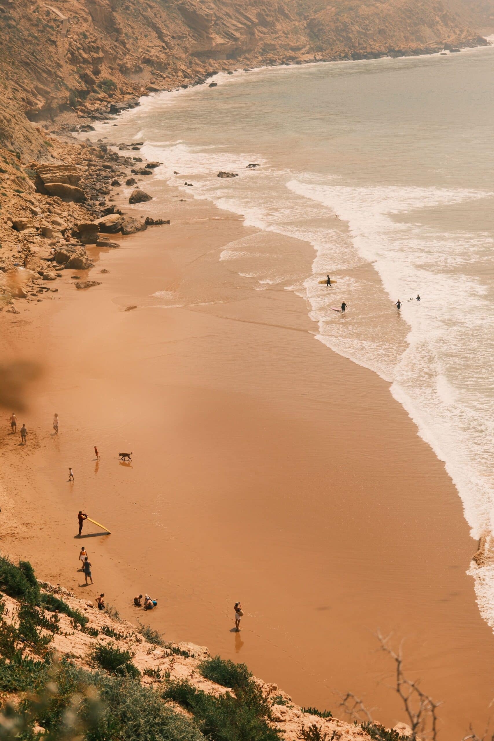 Vue aérienne d'une plage sauvage entourée de falaises au Maroc, avec des surfeurs entrant dans l'eau et des vacanciers sur le sable