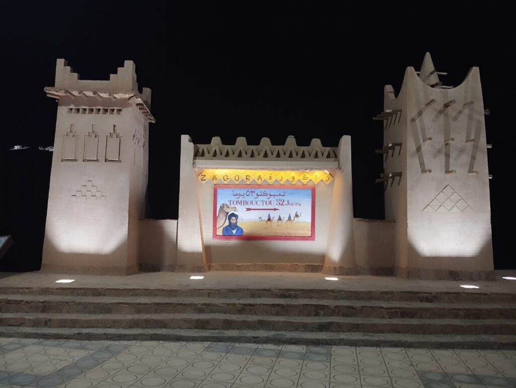 Porte de Zagora illuminée la nuit, avec le panneau historique "Tombouctou, 52 jours" à dos de dromadaire